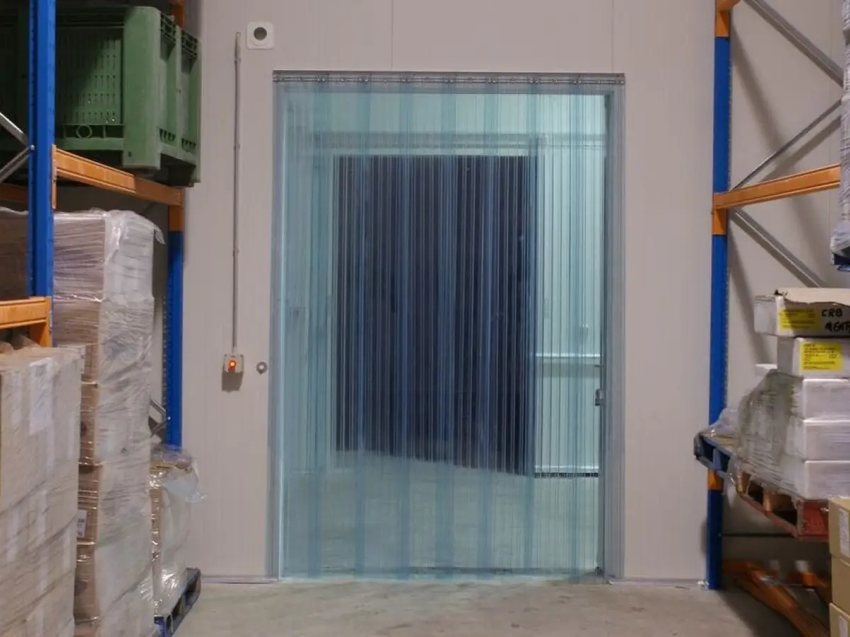 Strip curtains of PVC plastic sheets on an insulated freezer door frame within a food production & manufacturing warehouse.