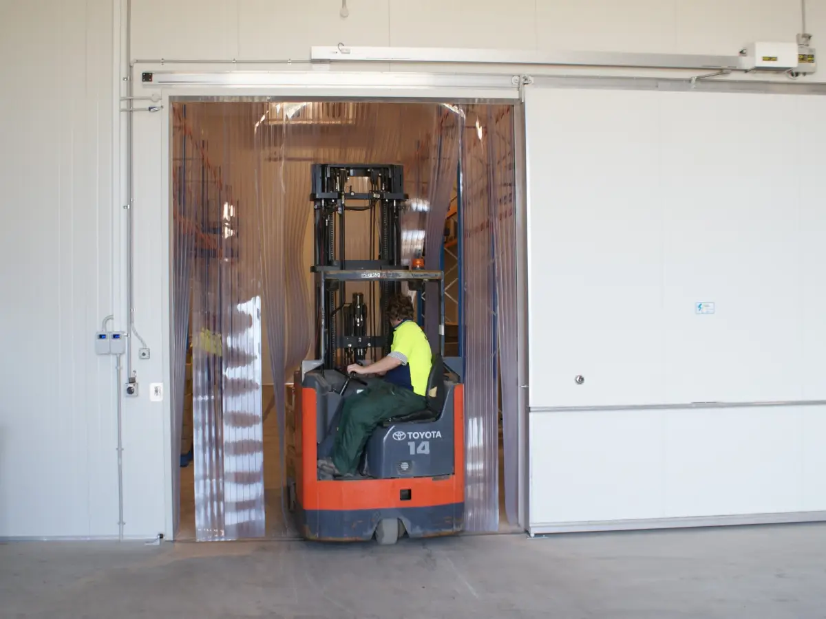 A forklift driving through an automatic sliding door with clear PVC strip curtain in an industrial factory.