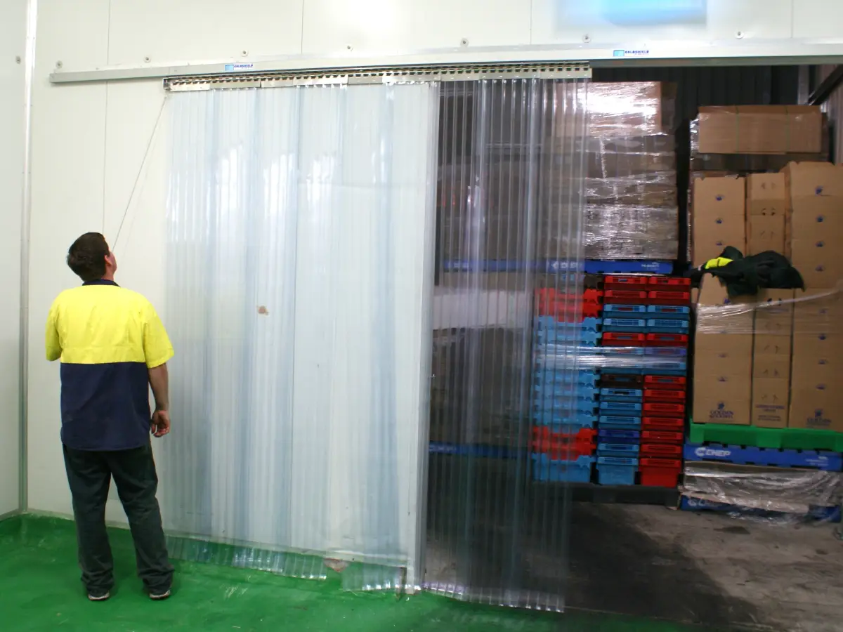 A factory employee opening clear plastic pvc strips on a thermal traffic door with sliding custom strip curtain mounting system in an industrial warehouse.
