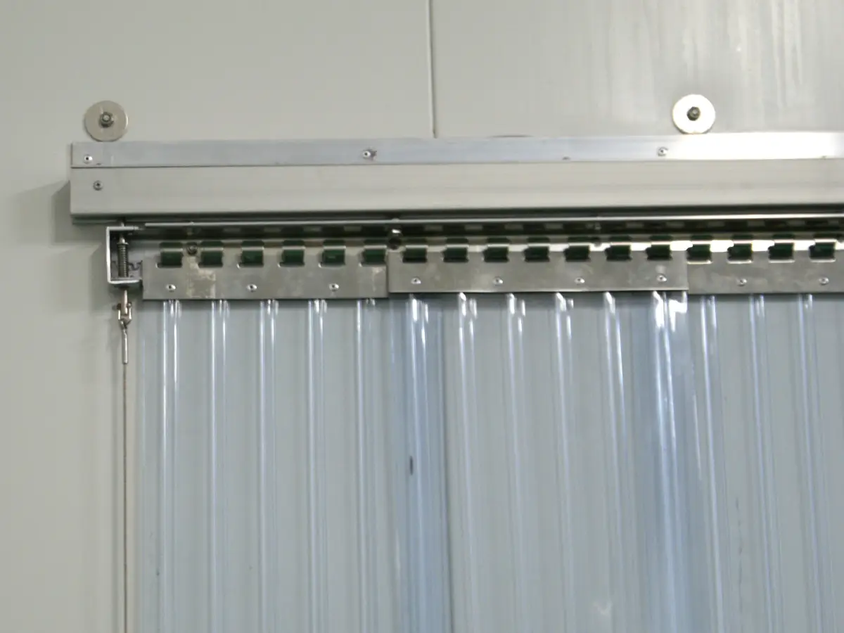 A sliding PVC strip curtain mounting system; clear plastic strips on hooks attached to a sliding cool room traffic door.